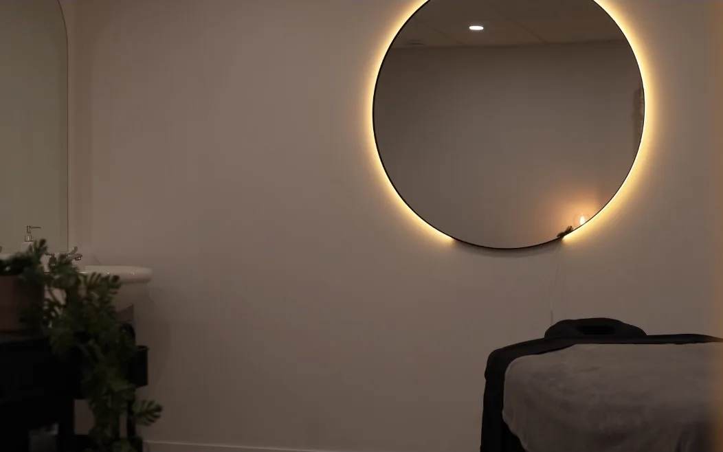La salle de soin : table de massage, miroir rond éclairé et plante verte